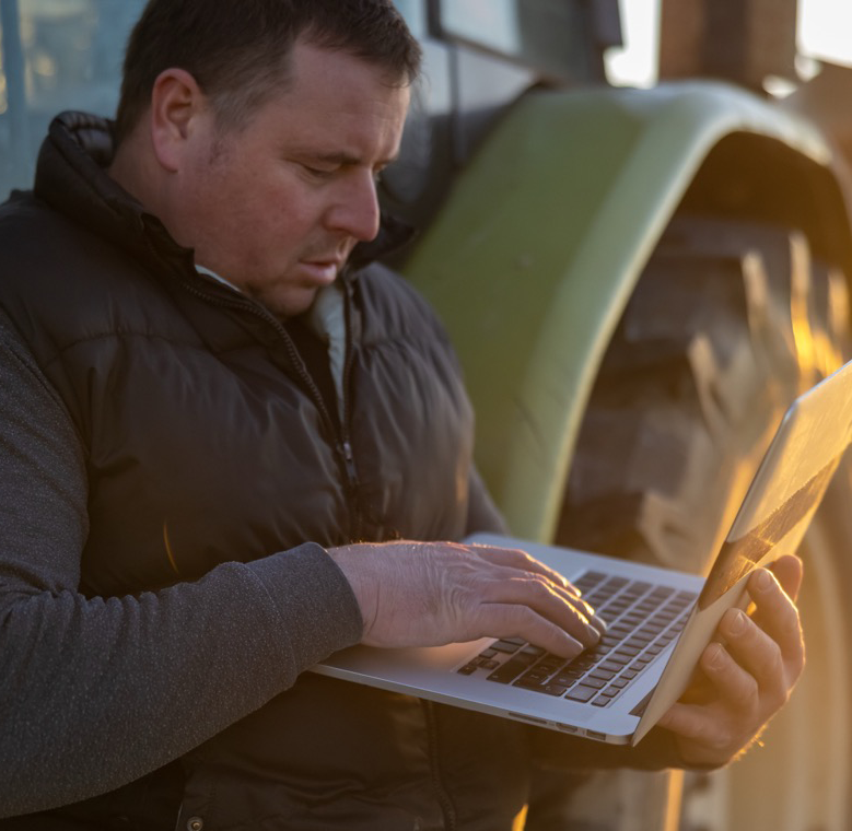 Man looking at laptop while resting against a tractor.