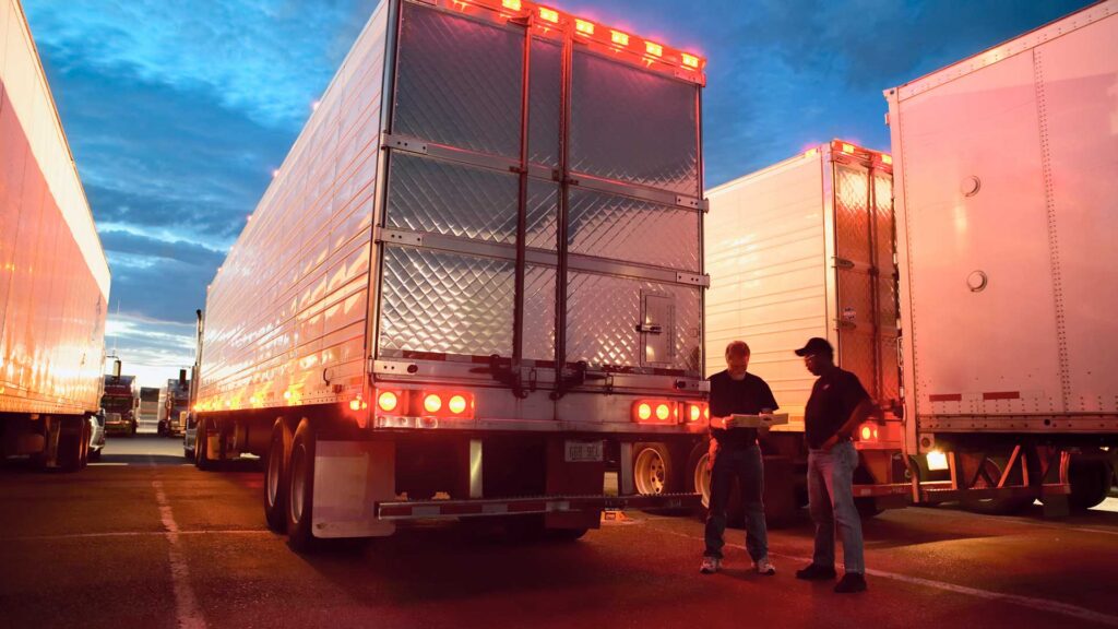 Two truck drivers checking dispatch papers while standing next to truck trailers in a large parking lot at night.