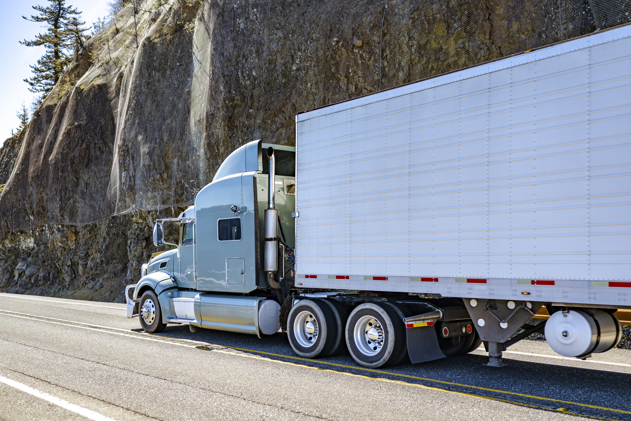 Industrial carrier gray big rig semi truck with extended cab for truck driver rest transporting cargo in refrigerator semi trailer running on mountain highway road with rock cliff in Columbia Gorge