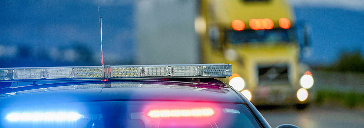 Police car with blue and red flashing lights in the foreground; a blurred yellow truck on a roadway behind it.