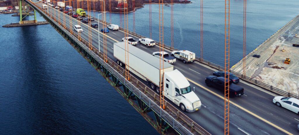 A busy bridge with a white semi-truck in a long line of vehicles crossing over a blue river or bay.