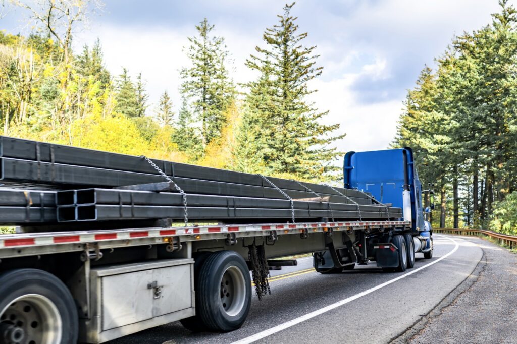 Blue semi-truck hauling long steel beams on a curved forest road.