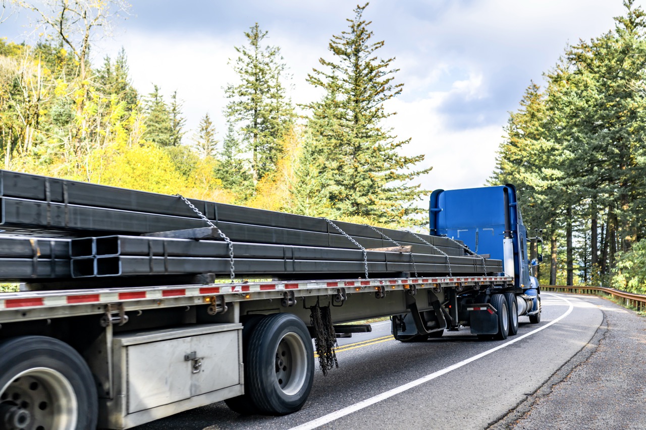 Blue semi-truck hauling long steel beams on a curved forest road.