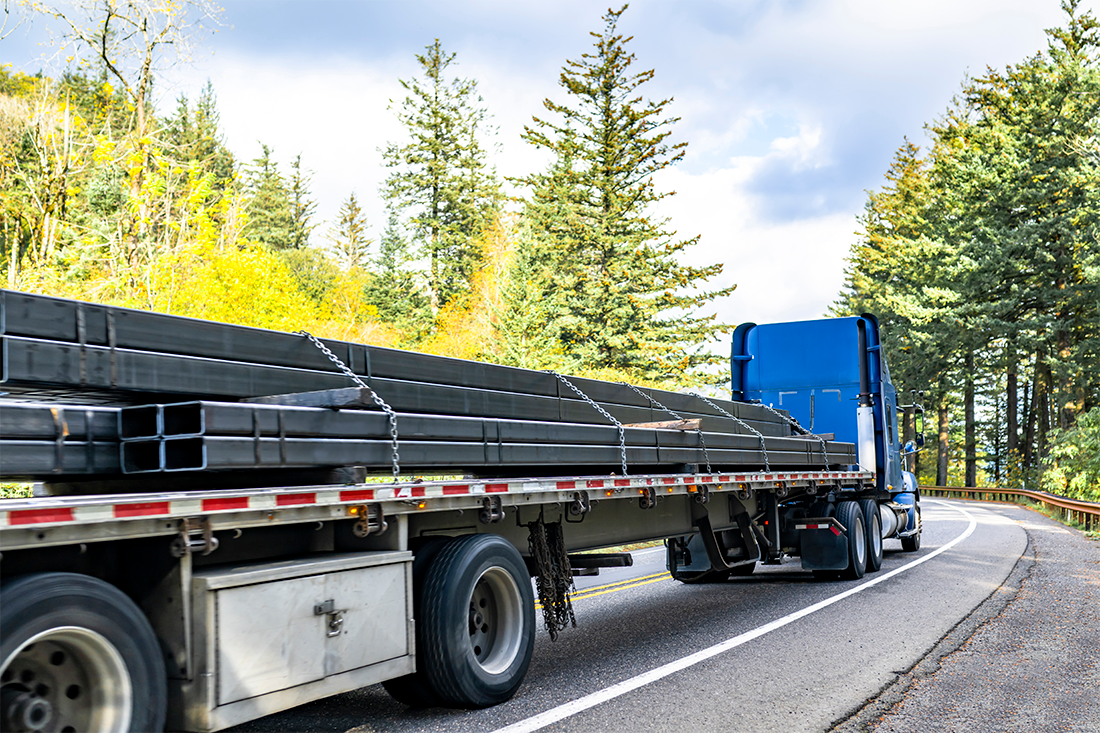 Semi-truck on a winding rural road carrying stacked steel beams secured with chains, with a forest of pines in the background.
