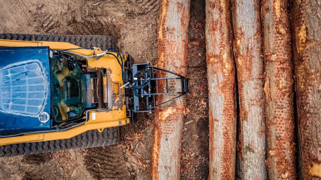 Top-down view of a yellow logging skidder beside piled logs in a bark-covered log yard.