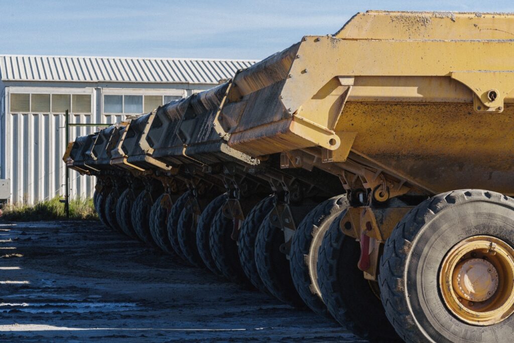 Row of large yellow dump trailer wheels and chassis, parked beside a corrugated metal building under a blue sky.