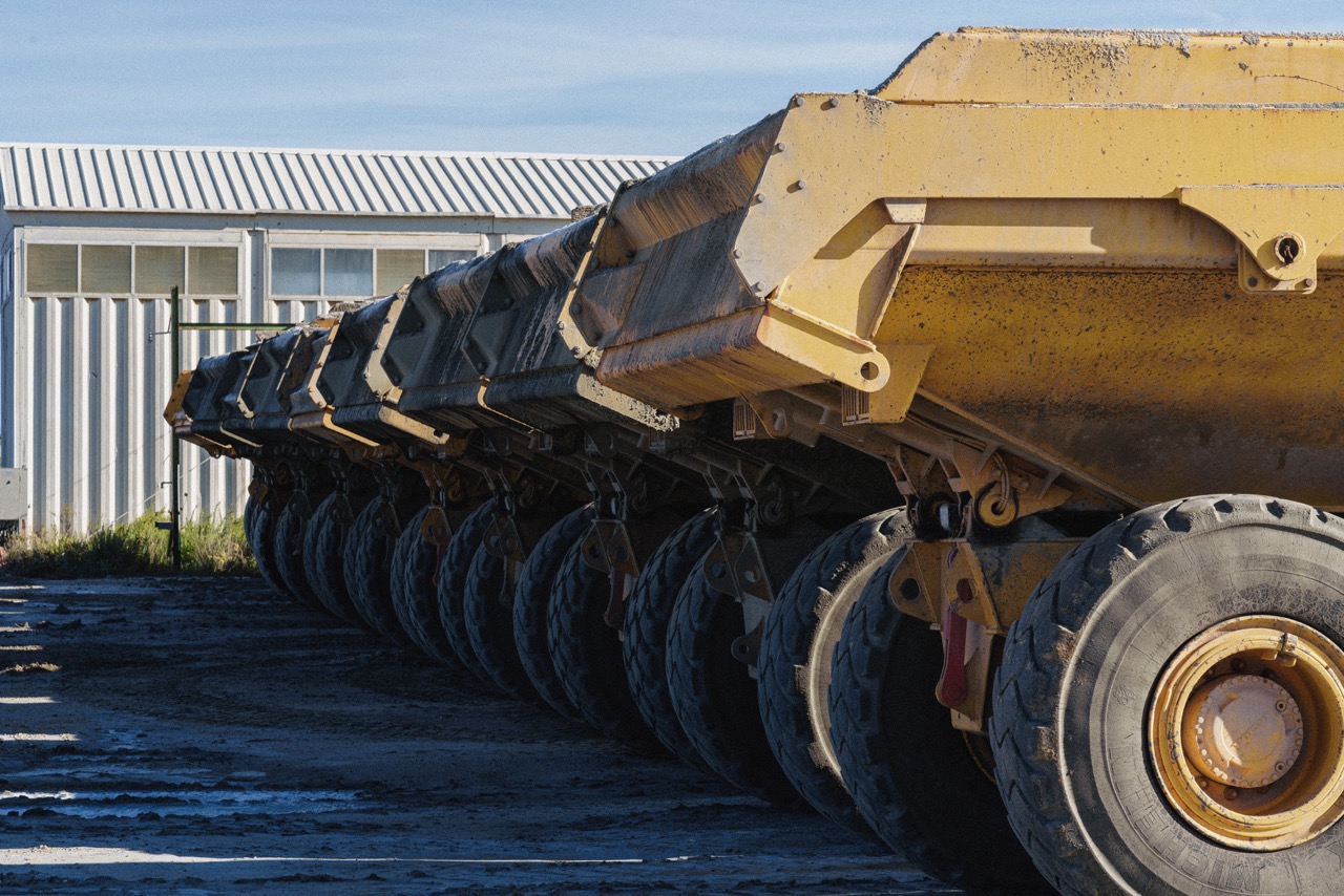 Row of large yellow dump trailer wheels and chassis, parked beside a corrugated metal building under a blue sky.