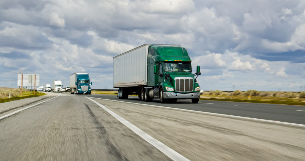 Green semi-truck with a large white trailer driving on a highway with other trucks in the distance under a cloudy sky