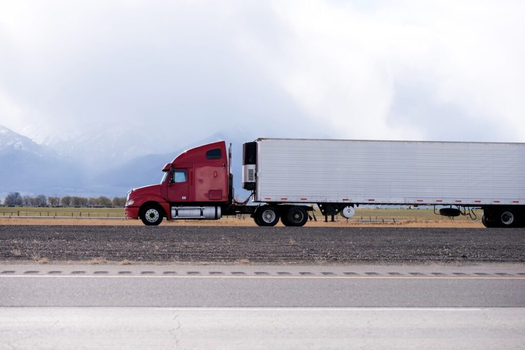 Red semi-truck with a long white trailer driving along a highway in a rural landscape with mountains in the distance.