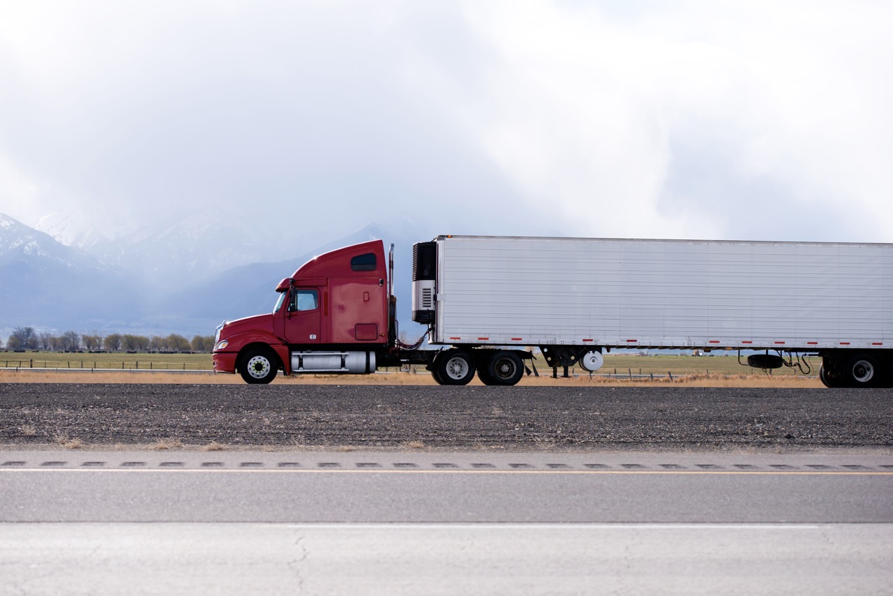 Red semi-truck with a long white trailer driving along a highway in a rural landscape with mountains in the distance.