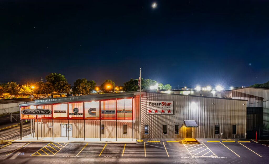 Night exterior of a freight service building with 'FourStar' and 'Freightliner' signage, illuminated parking lot beyond.