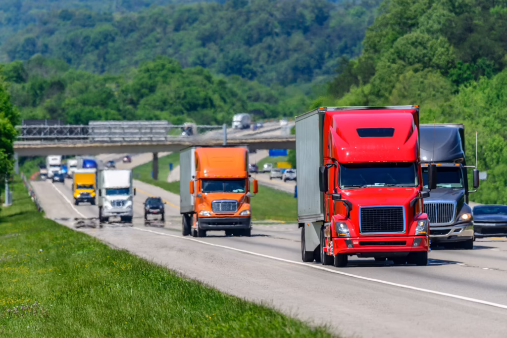 Red semi leading a line of trucks on a rural highway with green hills in the background, daytime