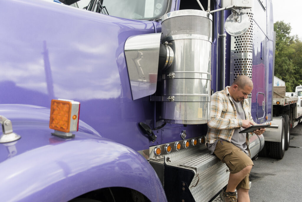 Man in a plaid shirt and shorts standing beside a large purple semi-truck, looking at a tablet in his hands.