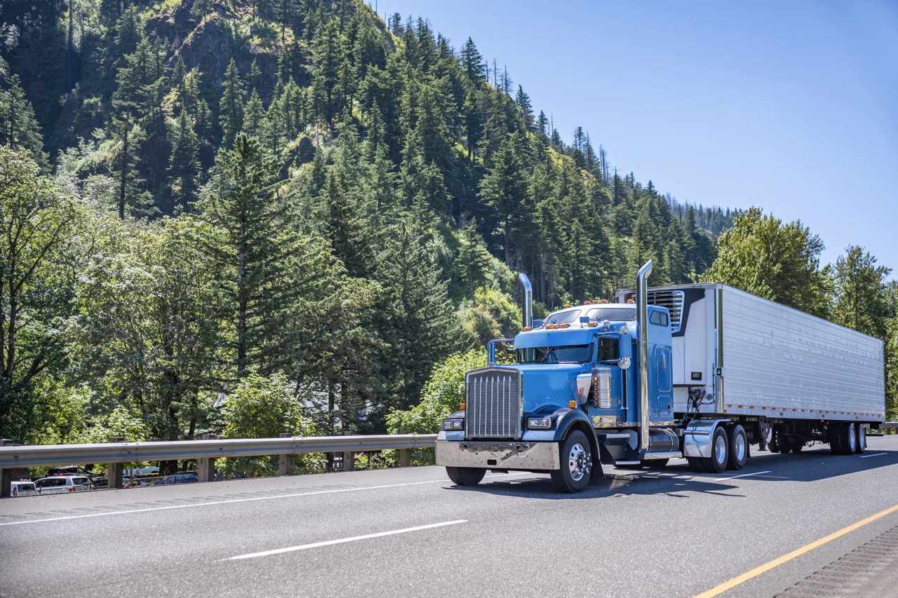 Blue semi-truck with a long trailer driving on a highway beside forested hills.