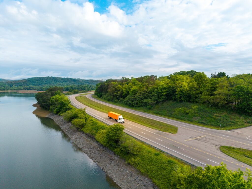Orange truck on a curved, divided highway beside a calm river, with green wooded hills and a cloudy sky in the background.