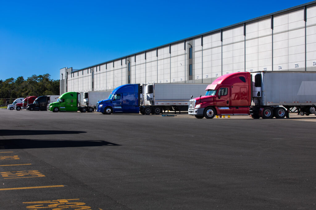 Row of colorful semi trucks parked beside a large industrial warehouse under a clear blue sky.