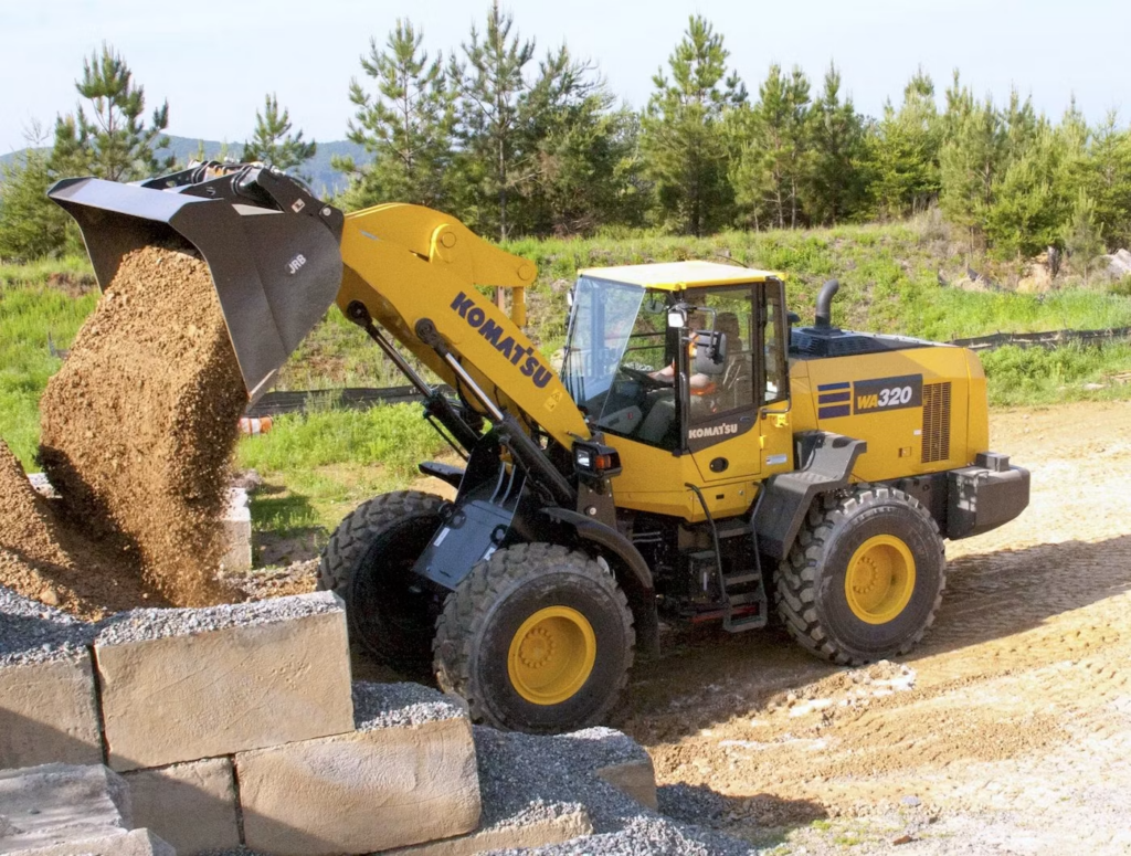 Yellow Komatsu WA320 wheel loader dumping soil into a pile at a construction site, with concrete blocks nearby on a dirt ground and trees in the background.